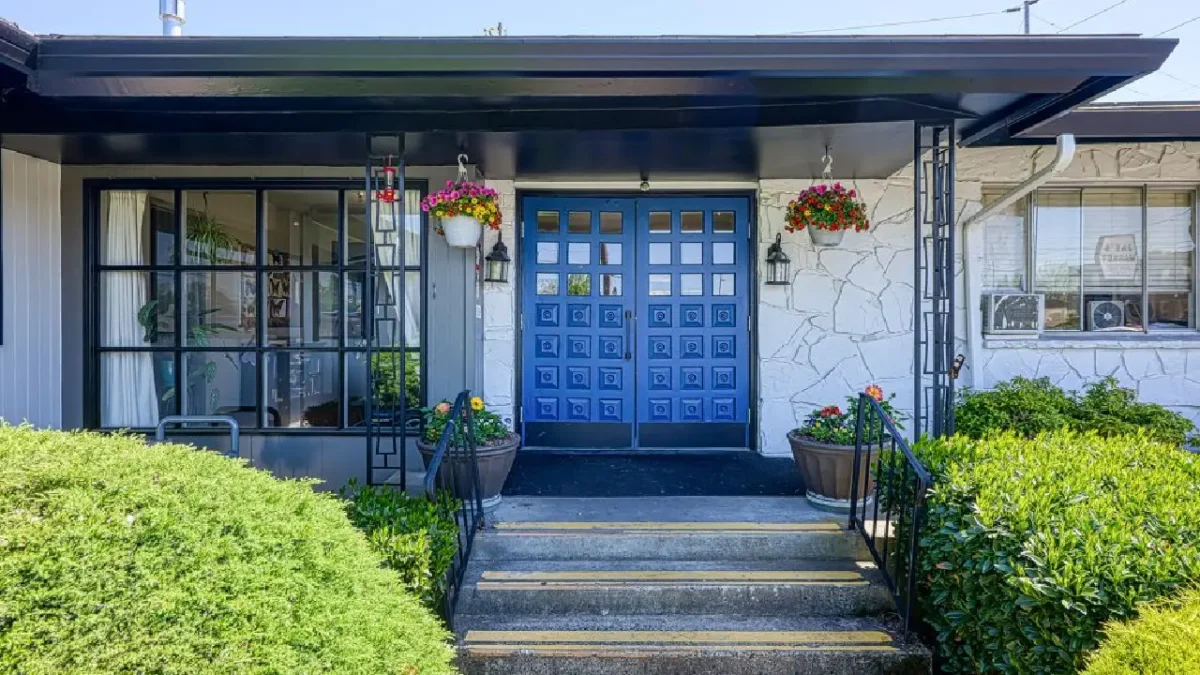 The front entrance of a white brick building with a black roof overhang, stairs, and a bright blue double door decorated with hanging flowers